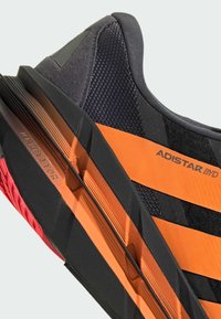 Black athletic shoe with orange stripes featuring a mesh and synthetic upper, textured sole, and graphic branding on the side.