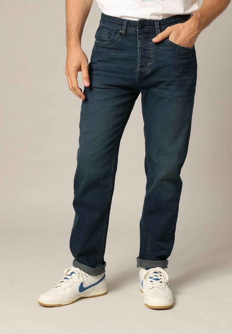 Jeans en denim bleu foncé avec une coupe droite, présentant cinq poches, une fermeture à bouton et un dégradé subtil. À associer avec des baskets blanches.