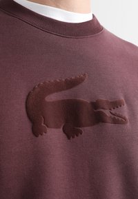 Felpa in pile bordeaux con un logo a forma di coccodrillo testurizzato, di un bordeaux scuro, sulla parte anteriore. Presenta un collo rotondo e un tessuto morbido.
