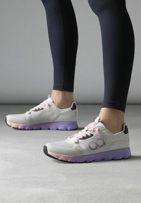 Scarpe atletiche grigio chiaro con suola viola, accenti di lacci rosa e un design in rete con un logo vicino alla caviglia. Suola in gomma testurizzata.
