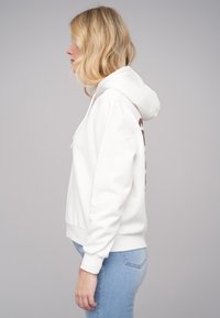 Hvit hettegenser med avslappet passform, myk tekstur, snorhette og mørkebrune grafiske detaljer på siden. Kombiner med lyse jeans.