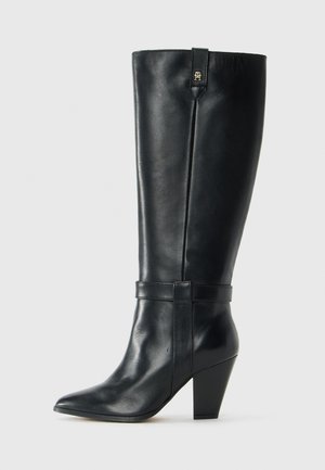 Black mid-calf leather boot with pointed toe, block heel, decorative strap around ankle, and small metal logo near top edge.