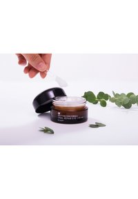 Pot noir de crème pour les yeux réparatrice à l'escargot avec un bord intérieur doré. Produit crémeux et blanc à l'intérieur, accompagné d'une petite spatule et de feuilles vertes.