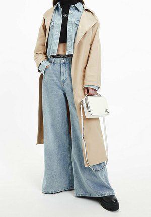 Mujer con abrigo tren beige, chaqueta vaquera azul claro y vaqueros de pierna ancha, sosteniendo un bolso blanco de Calvin Klein.