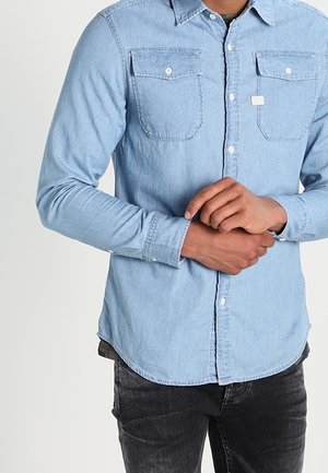 Homme portant une chemise en jean bleu clair avec des poches poitrine boutonnées et un jean noir, mains jointes à la taille.