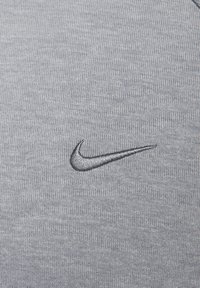 Szara tkanina z subtelną, dzianą fakturą, z ciemnoszarym haftowanym logo Nike w kształcie łyżwy umieszczonym po lewej stronie.