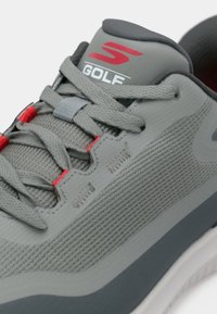 Sivá a červená golfová teniska s hornou časťou zo sieťoviny, sivými šnúrkami a červeným logom „S Golf“ na polstrovanom jazyku, detailným prešívaním a textúrovanou podrážkou.