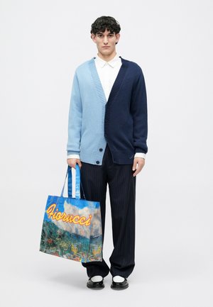 Tofarvet cardigan i lyseblå og marineblå, med V-udskæring og knapper. Modellen holder en blå tote bag med farverig grafik og "Fiorucci" tekst.