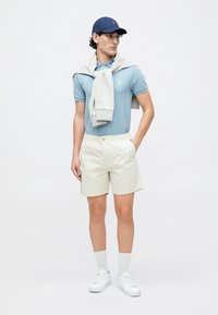 Light blue polo shirt, white shorts, gray sweater tied around the neck, navy cap, white sneakers, and crew-length socks, standing on a plain background.