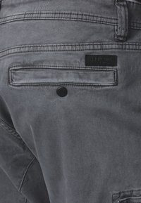 Gray denim pants featuring a back pocket with a black logo patch and a circular black button detail. Smooth texture with stitch accents.