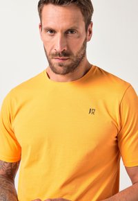 Krátký rukáv oranžové bavlněné tričko s malým černým logem na levé části hrudi a klasickým designem s kulatým výstřihem.