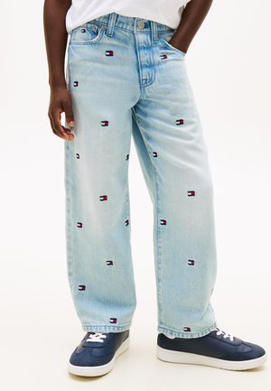 Jeans a gamba larga di colore azzurro chiaro con piccoli flag Tommy Hilfiger ricamati ovunque, indossati con sneakers blu navy e una maglietta bianca.