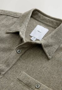 Woven shirt with a herringbone pattern in light brown and beige, featuring a collar, grey buttons, and a label indicating size M.