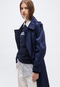 Νεαρή γυναίκα που φοράει στρωμένο ναυτικό μπλε παλτό τύπου trench, ναυτικό μπλε φούτερ με μερικά λευκά γράμματα, λευκό πουκάμισο με γιακά και λευκό παντελόνι με ναυτικές μπλε ρίγες.