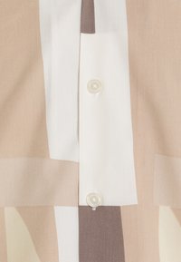 Camisa abotonada con una tela suave que presenta bloques de color geométricos en beige, crema y marrón claro, acentuada por un solo botón blanco.