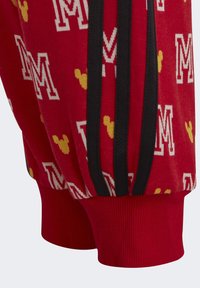 Pantalones deportivos rojos con patrones en blanco y amarillo de siluetas de "M" y de Mickey Mouse. Presentan franjas laterales negras y puños acanalados.