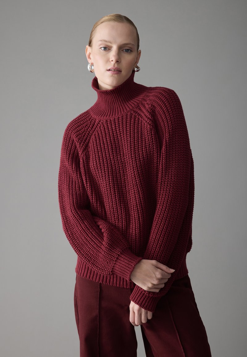 Bordeauxroter Strickpullover mit hohem Kragen, der über eine gerippte Textur und eine lockere Passform verfügt, kombiniert mit passenden burgunderfarbenen Hosen.