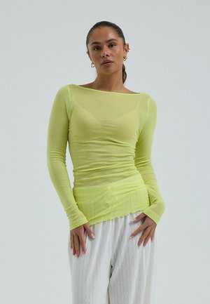 Femme aux cheveux foncés attachés en queue de cheval, portant un haut transparent vert citron à manches longues et un pantalon blanc ample à motifs, debout devant un fond uni.