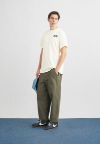 Krējuma krāsas kokvilnas T-krekls ar mazu logotipu, kombinēts ar brīvi pieguļošām olīvu zaļām biksēm. Melni sporta apavi ar baltām akcentiem. Nēsā zilu cepuri.