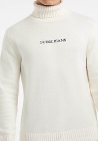 Maglione bianco lavorato a maglia con collo alto a coste, con logo nero "GUESS JEANS" sul petto e polsini e orlo a coste. Texture morbida.