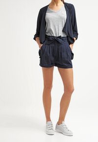Marinblå shorts med bälte i midjan, i kombination med en grå V-ringad t-shirt och en mörk kavaj. Avslutat med vita sneakers.