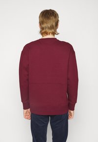 Felpa bordeaux con colletto a giro e polsini e orlo a coste, caratterizzata da un dettaglio con cucitura orizzontale sul retro. Tessuto in cotone morbido.