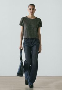 Młoda kobieta idąca w pomieszczeniu, ubrana w szary sweter z krótkim rękawem, szerokie ciemne dżinsy i niosąca czarną torebkę w lewej ręce.