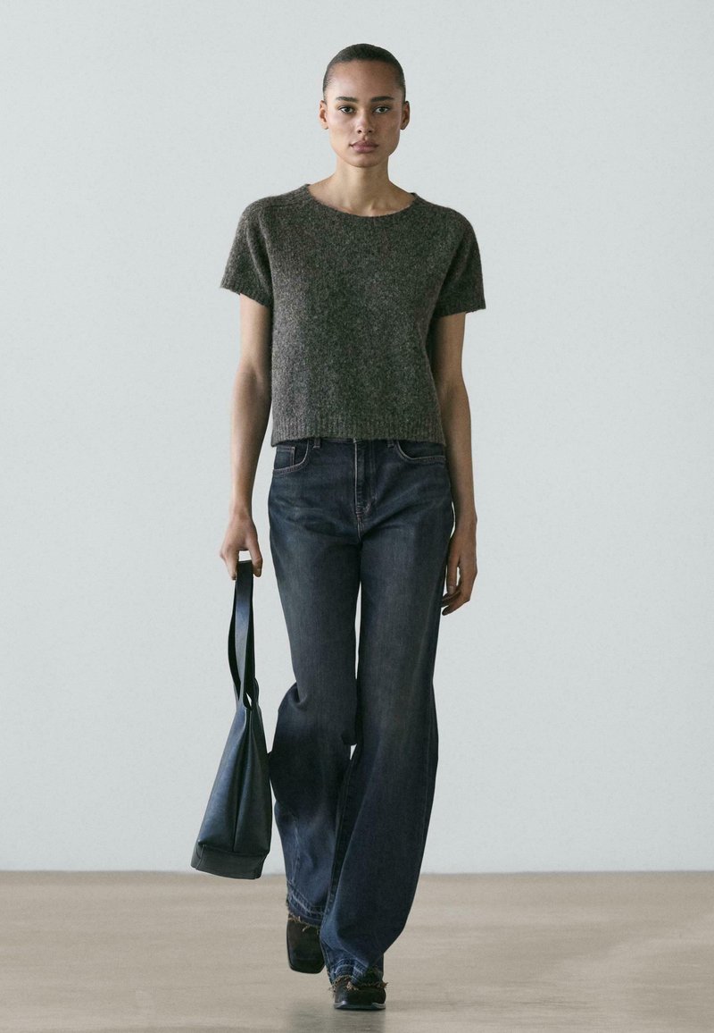 Młoda kobieta idąca w pomieszczeniu, ubrana w szary sweter z krótkim rękawem, szerokie ciemne dżinsy i niosąca czarną torebkę w lewej ręce.