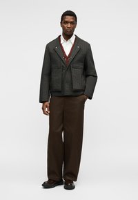 Homme portant une veste matelassée noire, une chemise blanche, un cardigan rouge, un pantalon marron à jambes larges et des chaussures foncées, debout devant un fond uni.