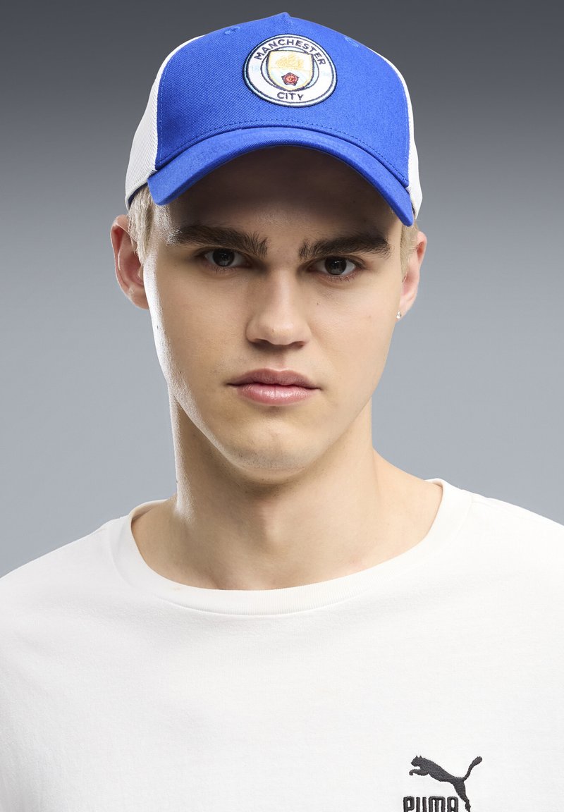 Jeune homme portant une casquette bleue et blanche de Manchester City et un t-shirt blanc Puma sur un fond gris.