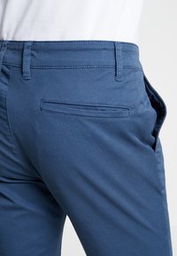 Blå bomullshängselbyxor med en slät textur, som har en bakficka och bältesögglor, visade ur en sidovinkel.