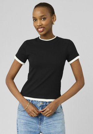 Jeune femme portant un haut noir à manches courtes avec bordure blanche et un jean bleu clair, souriante avec les mains jointes à la taille.