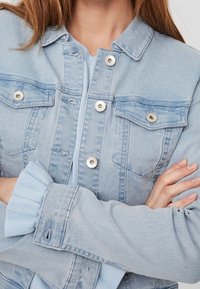 Veste en denim bleu clair avec boutons en métal argenté, deux poches poitrine et manches en tricot côtelé. Dispose d'un col classique et d'une fermeture à un bouton.