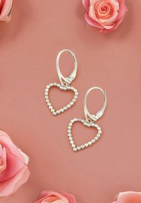 Silverfärgade hjärtformade örhängen med pärlliknande kulor, med en spänne med hävstång, mot en mjuk rosa bakgrund.