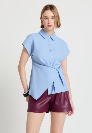 Femme porte un chemisier bleu clair à manches courtes avec boutons sur le devant et un détail noué, associé à un short en cuir rouge foncé et un bracelet noir.