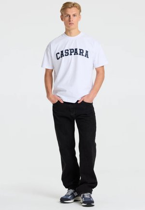 Ung mand stående med hænderne i lommerne, iført hvid t-shirt med teksten "CASPARA", sorte bukser og blå sneakers på ensfarvet baggrund.