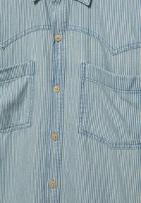 Chemise rayée bleu clair en denim avec deux poches avant, dotée de boutons marron et d'un col, présentant un tissu texturé.
