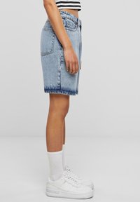 Kvinne i stripet crop top iført lyseblå denimshorts, hvite crew-sokker og hvite joggesko, stående på en ensfarget grå bakgrunn.