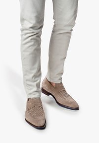 Mocassins en daim gris clair avec une texture tissée sur le bout. Les chaussures ont un petit talon et sont assorties à un pantalon slim de couleur claire.