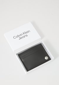 Portefeuille en cuir noir avec plusieurs emplacements pour cartes, présentant un accent en logo argenté. Emballé dans une boîte blanche portant l'étiquette "Calvin Klein Jeans".