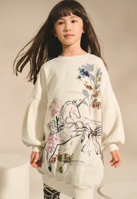 Hanorac alb cu mâneci supradimensionate, prezentând un grafic cu un unicorn, elemente florale și accente metalice. Textul de-a lungul marginii de jos spune „BUNĂTATE PENTRU TOȚI.”