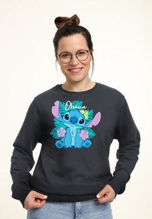 Frau mit Brille und Haarknoten trägt schwarzen Pullover mit blauem Cartooncharakter Stitch und tropischen Blumen, lächelt in die Kamera.