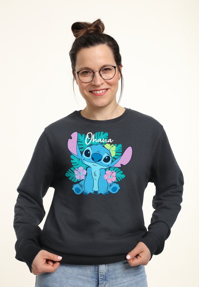 Kvinde med briller og håropsat hår iført sort sweatshirt med den blå tegneseriefigur Stitch og tropiske blomster, smilende mod kameraet.