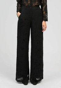 Femme portant un pantalon large en dentelle noir et un top en dentelle assorti, se tenant avec une main dans la poche devant un fond uni.