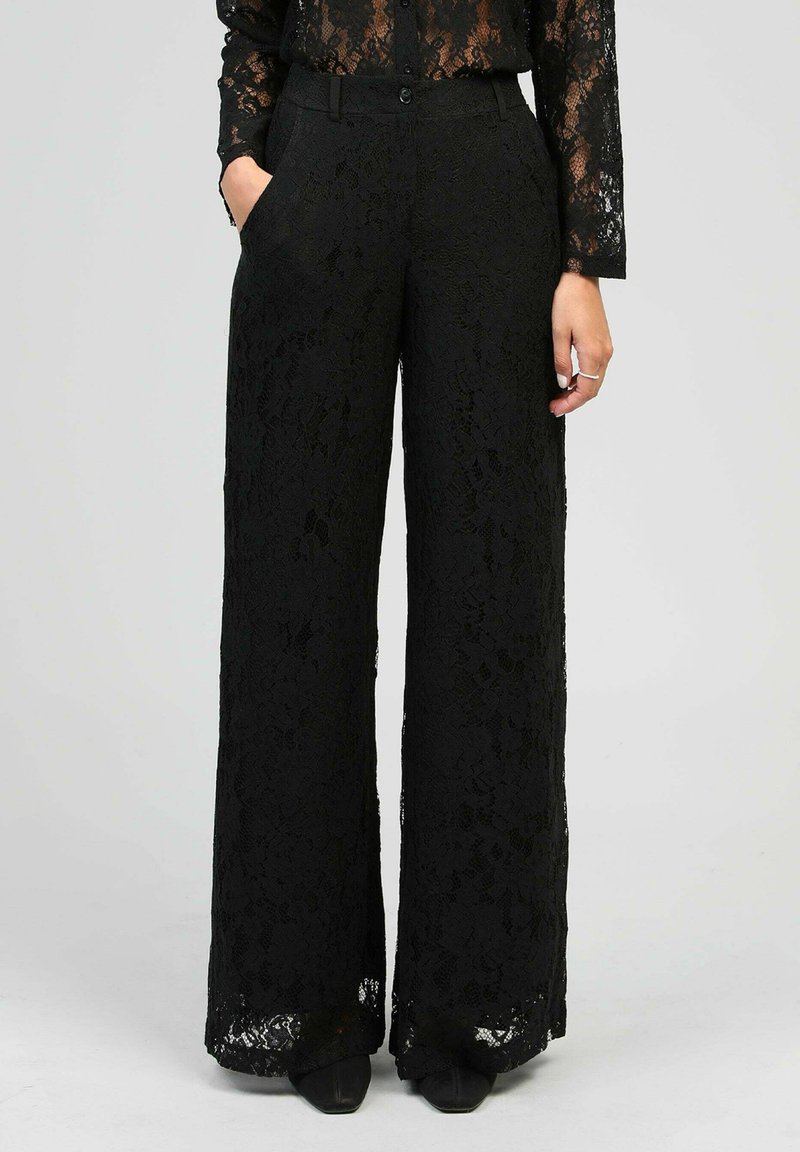 Femme portant un pantalon large en dentelle noir et un top en dentelle assorti, se tenant avec une main dans la poche devant un fond uni.