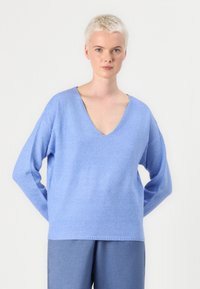 Pull en tricot bleu clair avec un profond col en V, des épaules tombantes et des manches longues. Texture douce, ourlet agrémenté de côtes, associé à un pantalon bleu.
