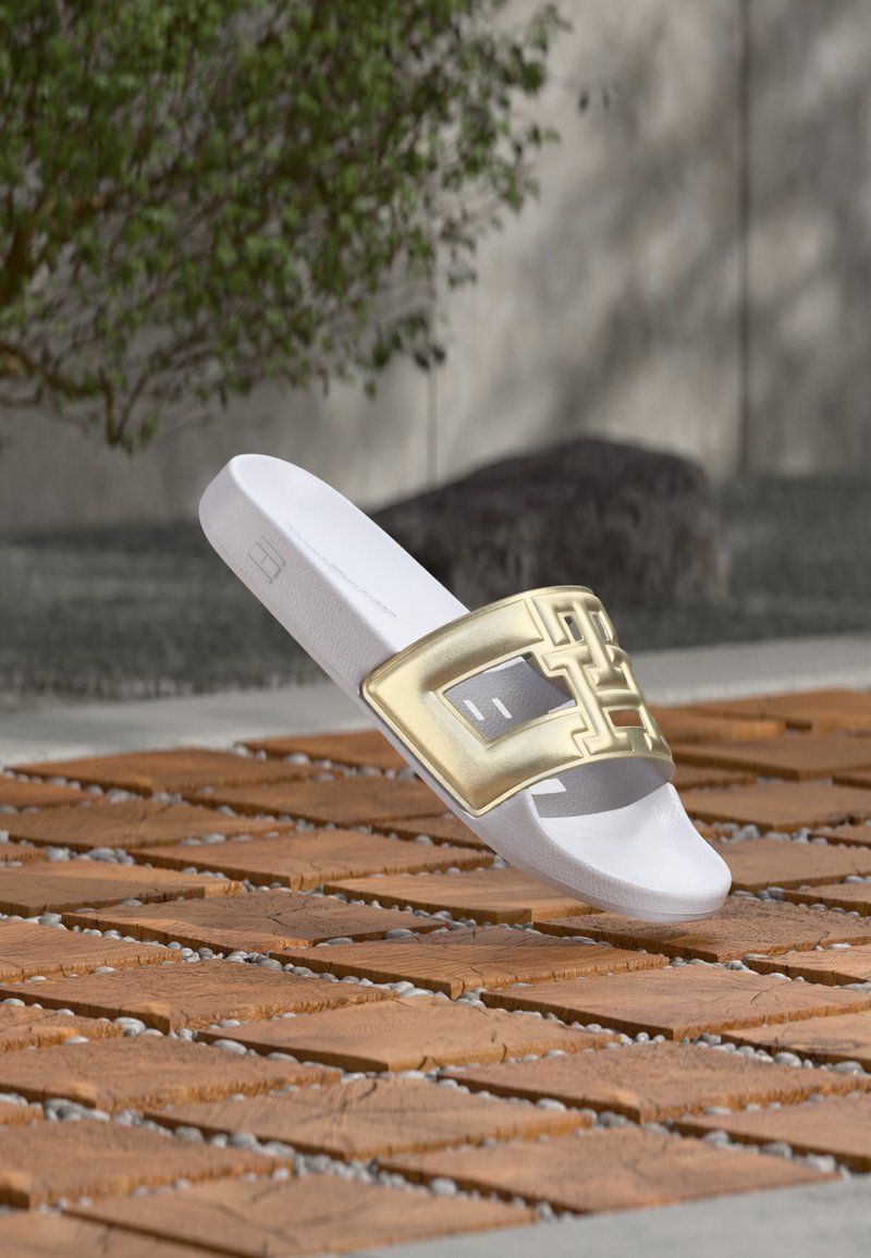 Sandale blanche à enfiler avec une large bride géométrique métallique dorée reposant sur une surface extérieure en bois à lattes avec de la verdure en arrière-plan.