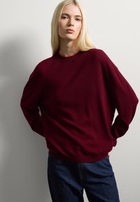 Femme blonde portant un pull maroon ample et un jean bleu foncé, se tenant les mains derrière le dos contre un mur blanc.