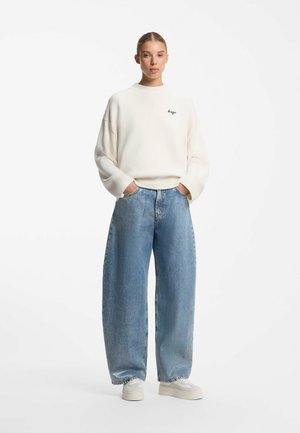 Ung kvinde iført en hvid sweater med teksten "hugo", brede lyseblå jeans og hvide sneakers på hvid baggrund.