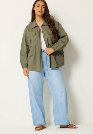 Mujer con chaqueta verde oliva de gran tamaño, top blanco, vaqueros de pierna ancha azul claro y sandalias marrones, de pie frente a un fondo claro y liso.